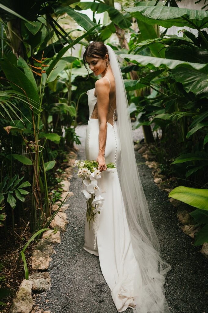Bride looking down at her white tropical bridal bouquet standing in the Tulum jungle. 