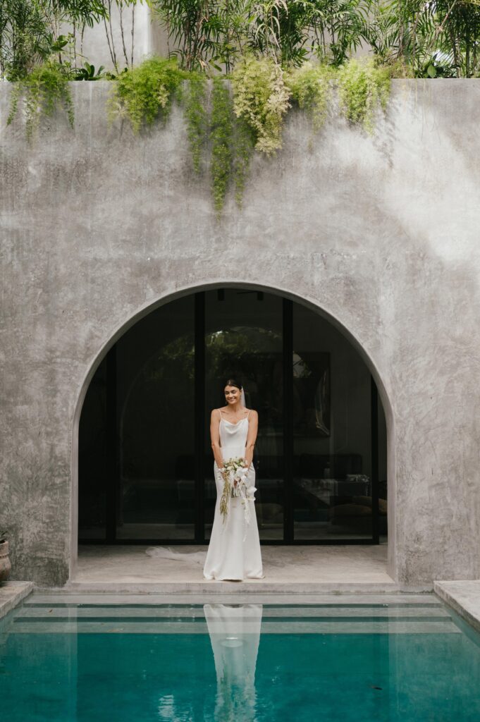 Bride standing under archway by swimming pool villa in aldea zama tulum just after wedding ceremony. 