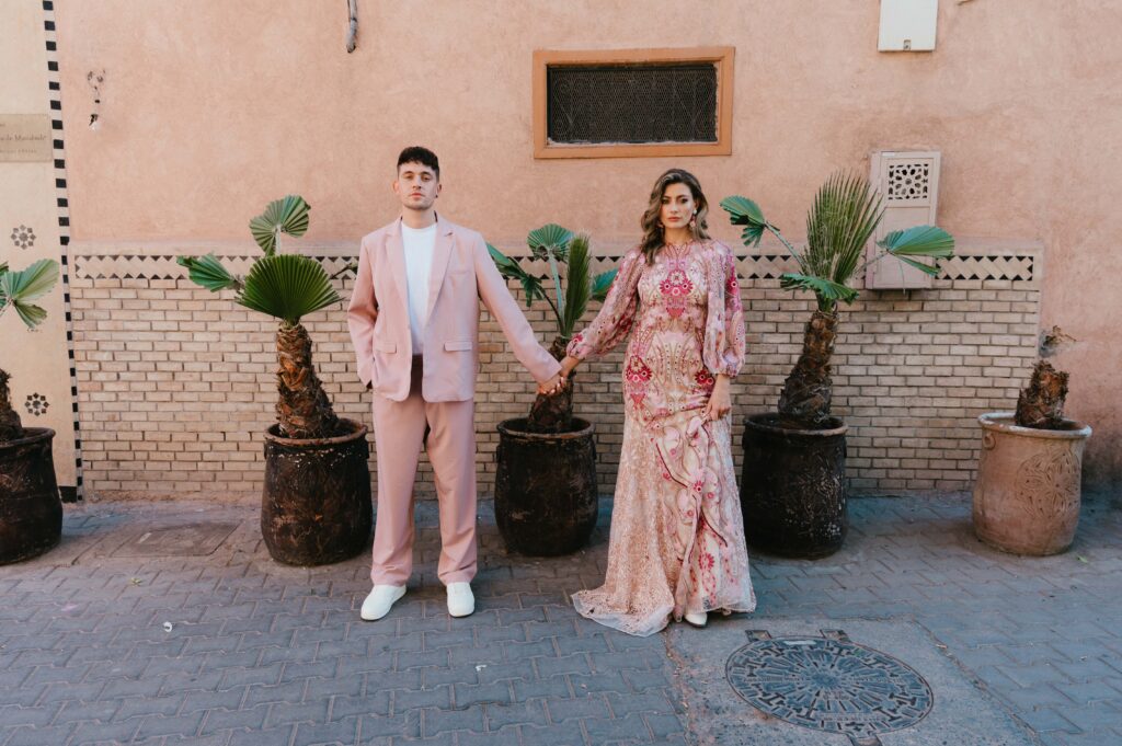 Wide angle of Lisa and Paul, in wedding attire, holding hands in front of cactus pots and pink wall in the Marrakech Medina. 
