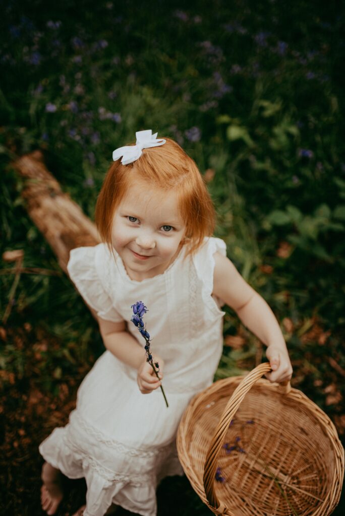 Little red hair girl looking up at the camera, holding a wicker basket in one hand and a bluebell in the other hand. She is sitting on a log in the bluebells. 