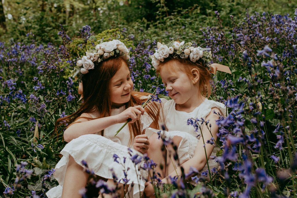Two sisters with matching ginger hair, wearing Zara white dresses and white flower crowns, sitting in the bluebell woods. 