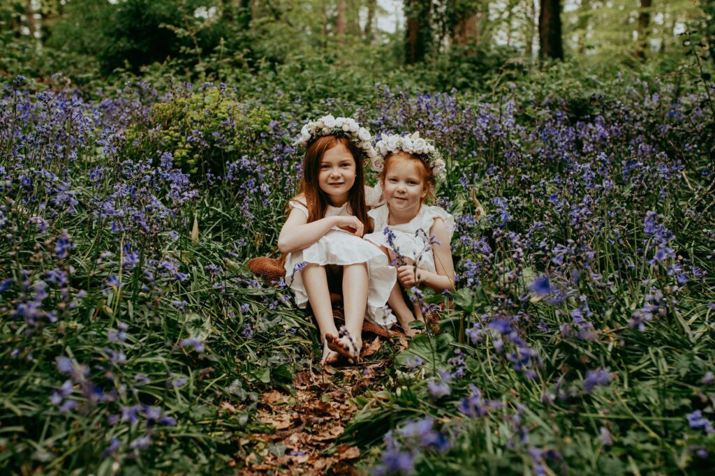 Two sisters with matching ginger hair, wearing Zara white dresses and white flower crowns, sitting in the bluebell woods. 