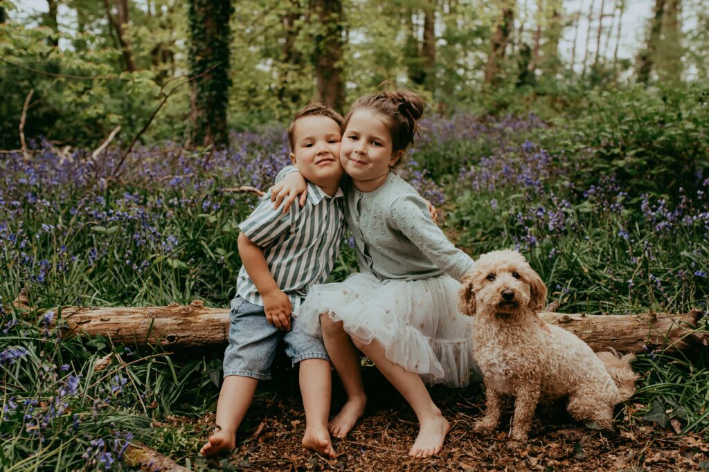 Arlo and Ivy hugging their dog Cooper and sitting on a log in the bluebells. 