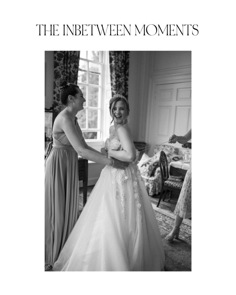 Candid photo in black and white of Amanda smiling at the camera while being helped in her wedding dress by her mother and maid of honour in the bridal suite at Kingston Bagpuize House.