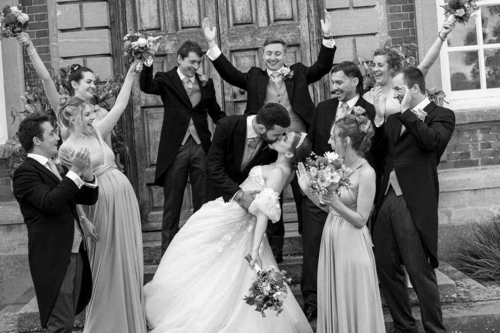 A joyful and candid wedding photo in black-and-white of a bride and groom kissing in a playful dip pose, surrounded by their bridal party celebrating with raised arms, smiles, and laughter in front of a grand wooden door.