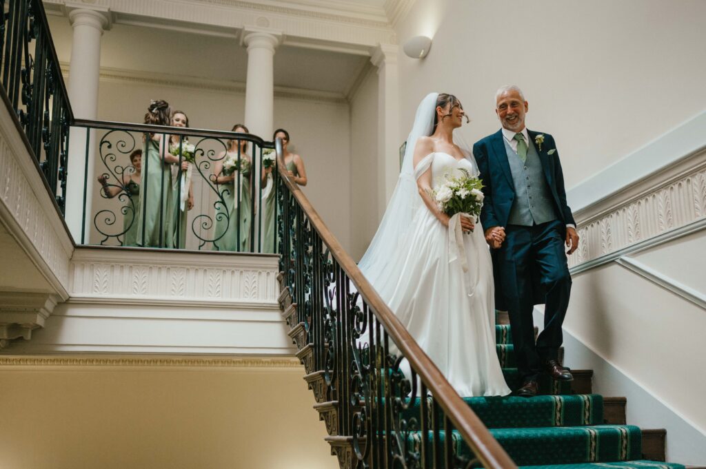 Portrait of bride and her father walking down the stairs with bridesmaids waiting in the background.