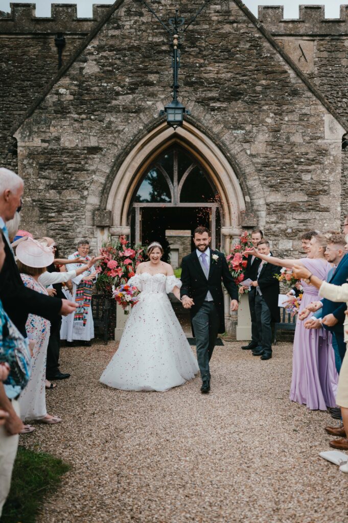 Amanda and John surrounded by guests as they are showered with colorful confetti outside St. Mary the Virgin Church after their ceremony.