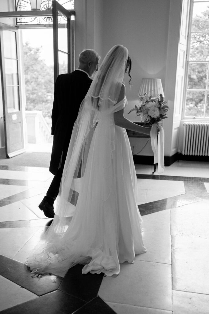 Black and white portrait of the bride and father of the bride in the entrance at Botleys Mansion. Bride is holding a bouquet and they are both facing away from the camera.