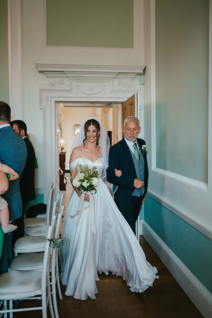 The bride, Charlie, is in a white dress holding a bouquet walks with her father in the ceremony room at Botleys Mansion.