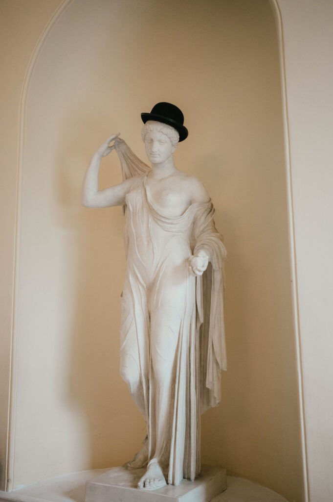 A classical marble statue draped in fabric, adorned with a modern black bowler hat, standing in an arched alcove with a beige background.