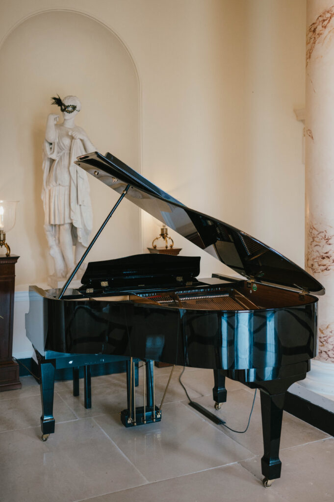 A sleek black grand piano with its lid open, set in an elegant room with cream walls and a marble column. In the background, a marble statue wearing a masquerade-style mask adds a playful and artistic touch.