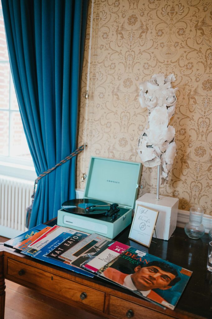 A vintage Crosley record player in mint green sits on a wooden desk, surrounded by an assortment of colorful vinyl records. Behind it, a textured plaster sculpture and a framed sign that reads "Old Time Records" enhance the retro aesthetic, with vibrant blue curtains and patterned wallpaper completing the scene.