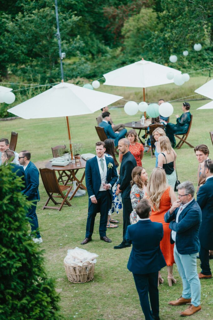 At Charlie and Martin's wedding at Botleys Mansion, a group of people socialises outdoors before the ceremony, with umbrellas, tables, and chairs elegantly set on the grassy area.