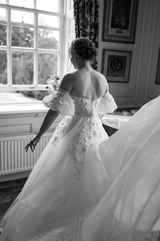 Amanda standing by the window in her wedding dress, having a quiet, reflective moment before the ceremony, bathed in soft natural light.