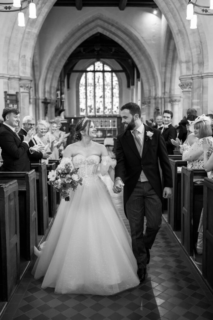 Amanda and John walking down the aisle at St. Mary the Virgin Church in Buckland, with smiles and excitement on their faces.