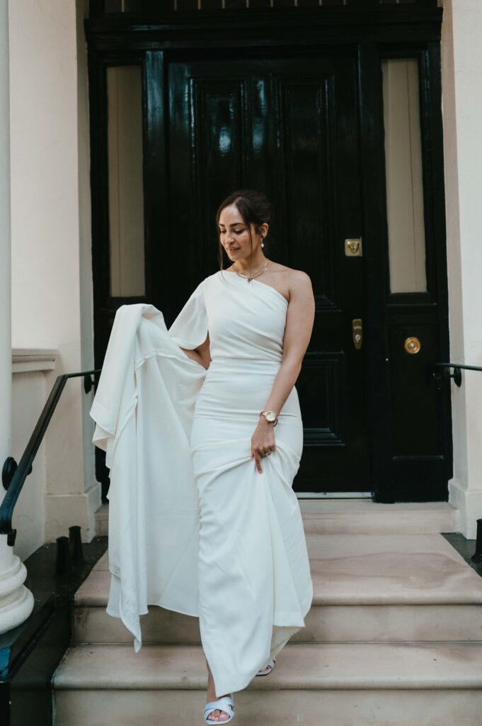 Raissa radiates grace and beauty as she stands on the steps of a classic London townhouse, her contemporary white wedding gown highlighted by the bold black door and stately columns that frame the scene.