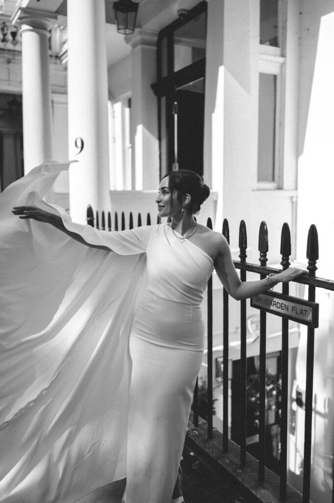 Captured in classic black and white, Raissa lifts the delicate fabric of her gown with grace, the interplay of light and shadow accentuating her poised elegance against the vintage charm of the townhouse backdrop.