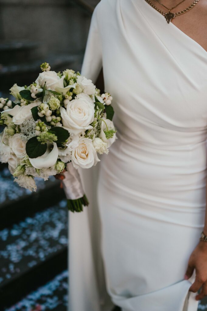 Close-up of a bride holding a delicate bouquet of white roses, calla lilies, and greenery, with the elegant details of her modern white gown and layered necklaces visible.