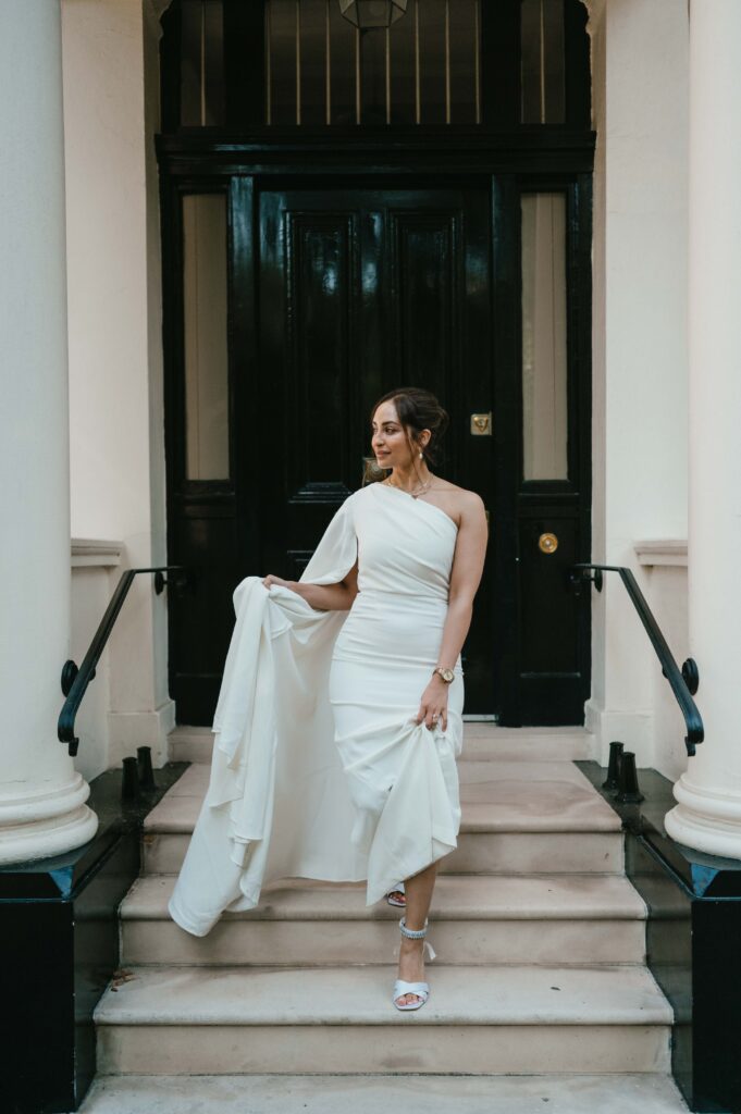 Raissa stands gracefully on the steps of an elegant townhouse in her modern white wedding gown, her soft smile radiating confidence and beauty as the black door and classic columns frame her.