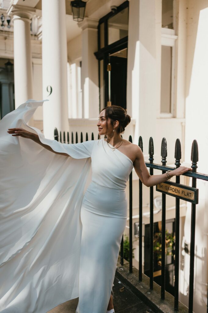 Raissa playfully lifts the flowing fabric of her wedding dress against the backdrop of a charming townhouse, her expression serene and radiant as sunlight filters through, highlighting her timeless elegance.