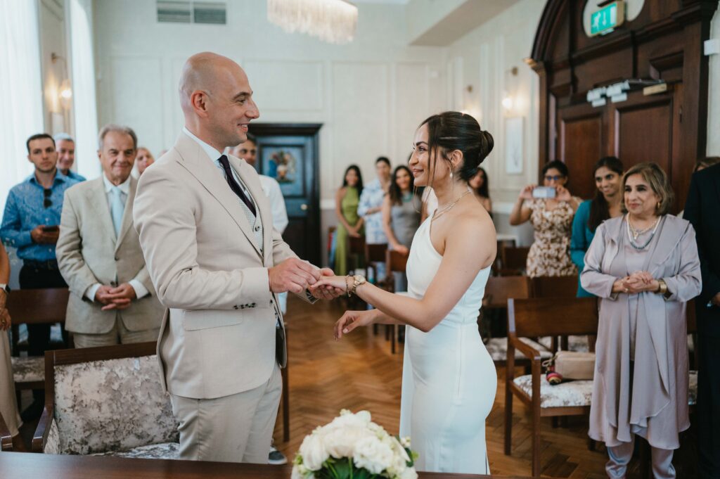 A romantic and modern wedding ceremony inside Chelsea Old Town Hall, featuring the couple exchanging rings, surrounded by soft natural light, elegant wooden paneling, and their closest family and friends seated in anticipation.