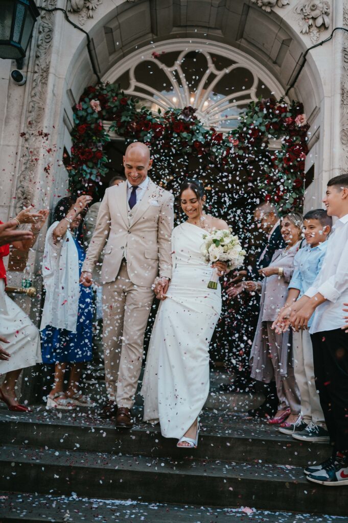 Raissa and Vasileios exit Chelsea Old Town Hall, hand in hand, as guests throw confetti in celebration.