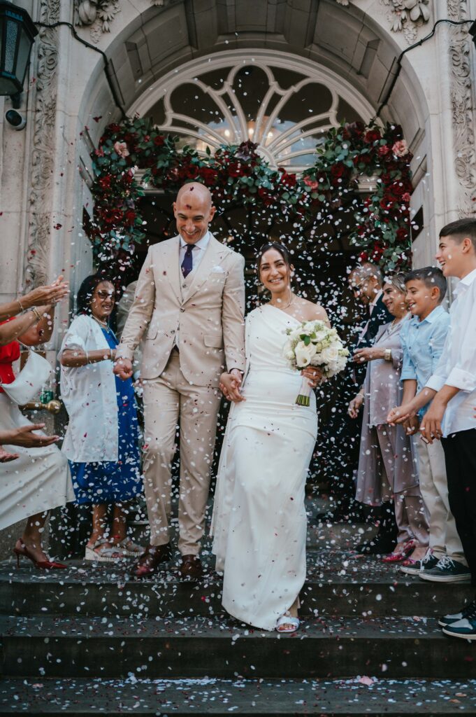 Raissa and Vasileios step out of Chelsea Old Town Hall under a cascade of confetti, surrounded by family and friends. The bride stuns in a chic one-shoulder cream dress by Solace London, while the groom beams in a beige suit, celebrating their modern and intimate wedding day in the heart of London.