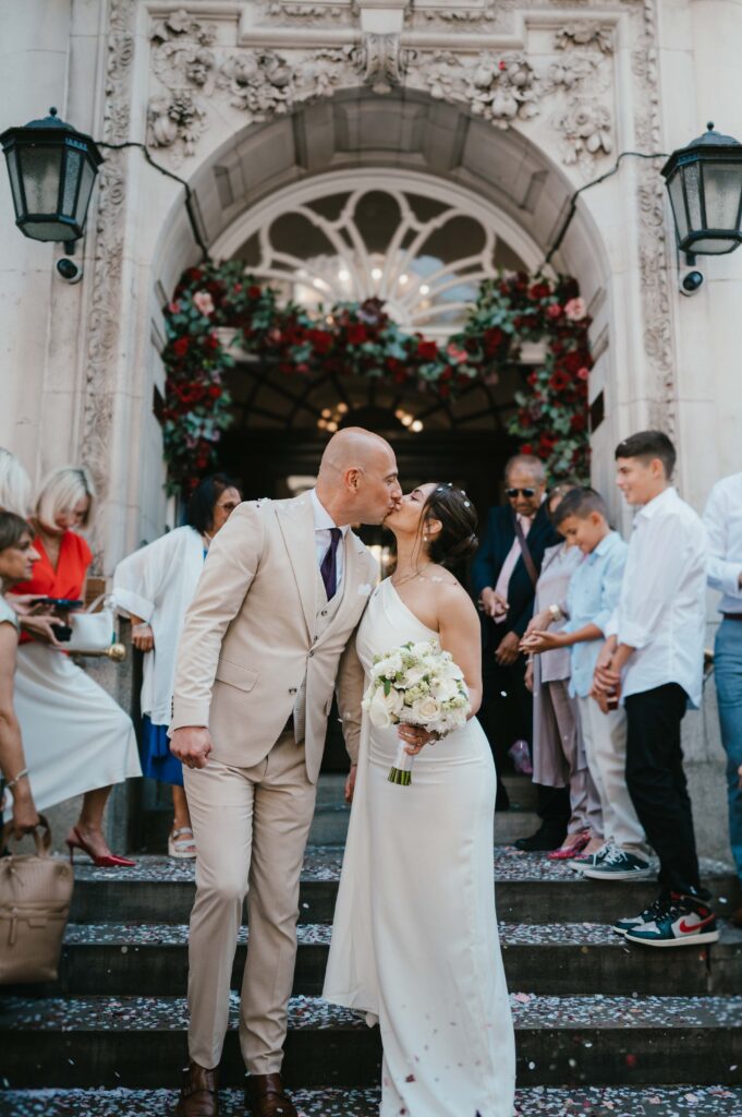 The bride and groom shares a kiss on the steps of Chelsea Old Town Hall, surrounded by friends and family.