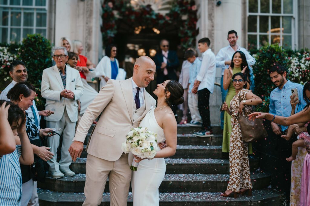 Raissa and Vasileios step out of Chelsea Old Town Hall under a cascade of confetti, surrounded by family and friends. The bride stuns in a chic one-shoulder cream dress by Solace London, while the groom beams in a beige suit, celebrating their modern and intimate wedding day in the heart of London.