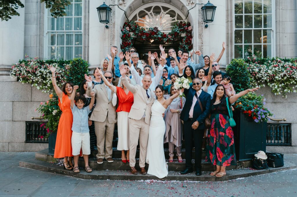 Family photo of the bride and groom surrounded by all their guests and cheering with their arms up in the air. 