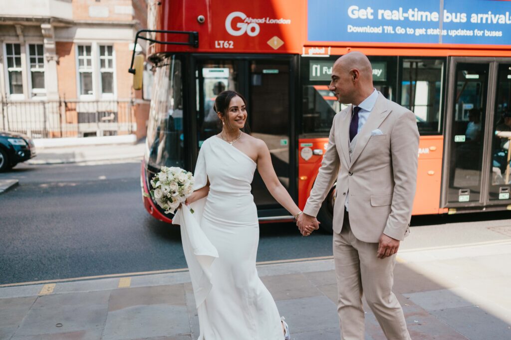 Raissa and Vasileios stand happily in front of a classic London red double-decker bus, smiling at the camera as they pause for a moment during their wedding celebration.