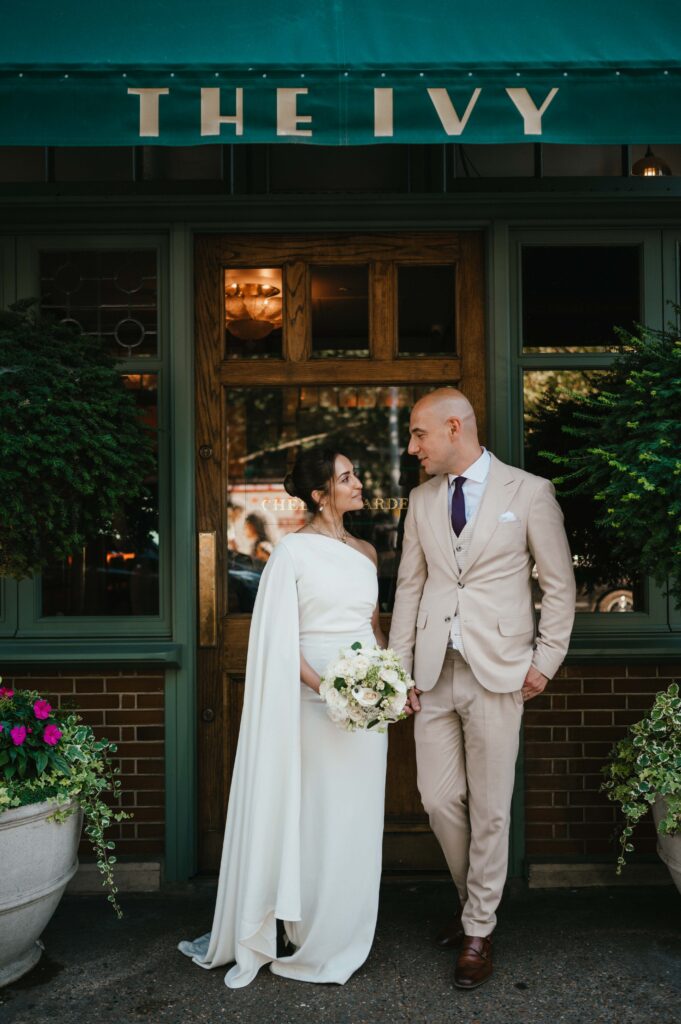 Raissa and Vasileios pose outside The Ivy, their wedding venue, with Raissa in her elegant bridal gown and Vasileios in a sharp suit, standing together in front of the restaurant's iconic entrance.