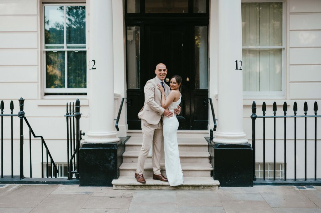 Raissa and Vasileios pose in front of a classic white townhouse at door number 12, standing on the steps with elegant black railings on either side. The groom, dressed in a beige suit with a tie, embraces the bride, who stuns in a sleek, one-shoulder white gown. Both are smiling warmly, radiating joy on their wedding day in London.