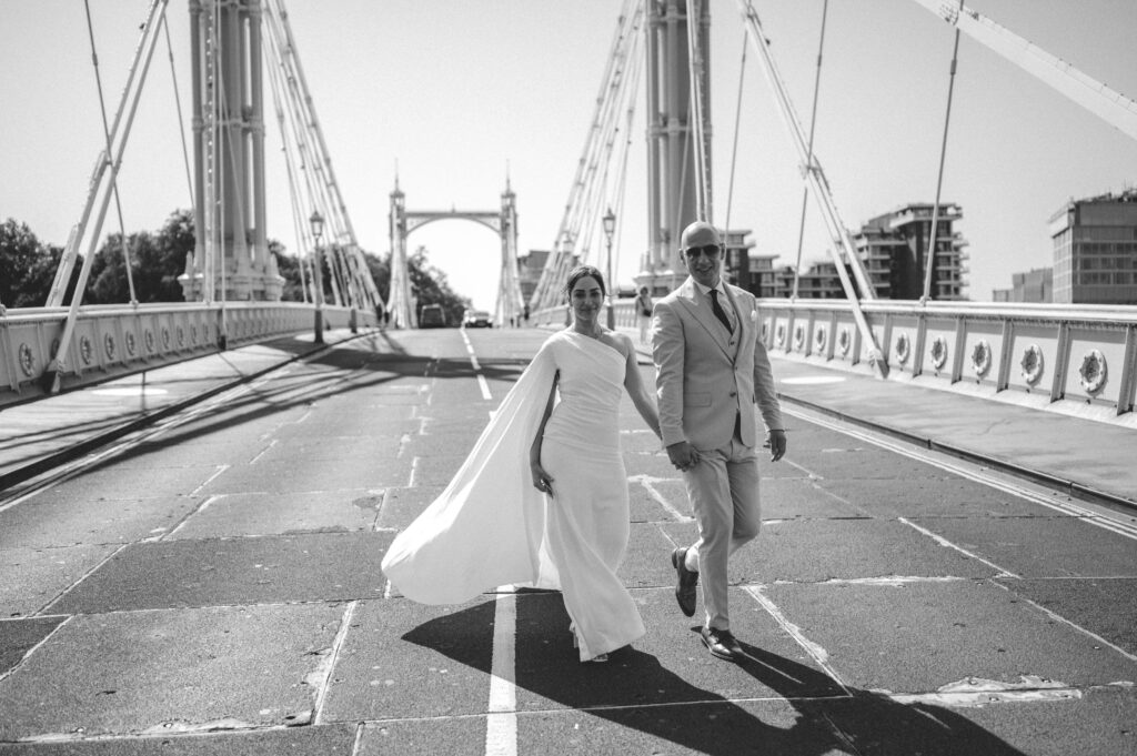 The couple walks hand-in-hand along Albert Bridge, its delicate white and pastel arches illuminated by the warm summer sun.