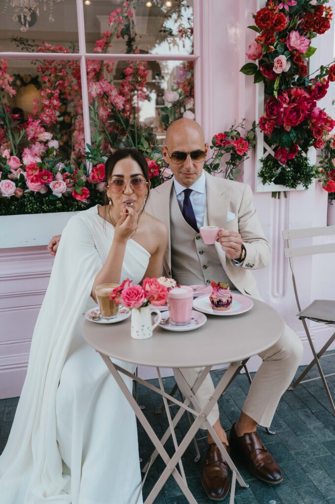 The couple shares a lighthearted moment at Peggy Porschen Café, with Raissa taking a bite of her cupcake while surrounded by the café's iconic pink floral backdrop.