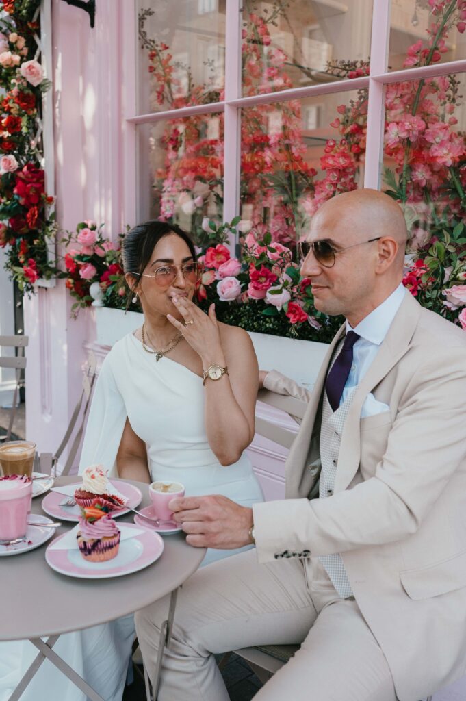 The couple enjoys a lighthearted moment with pastel-colored cupcakes in hand, surrounded by the café’s iconic floral décor.