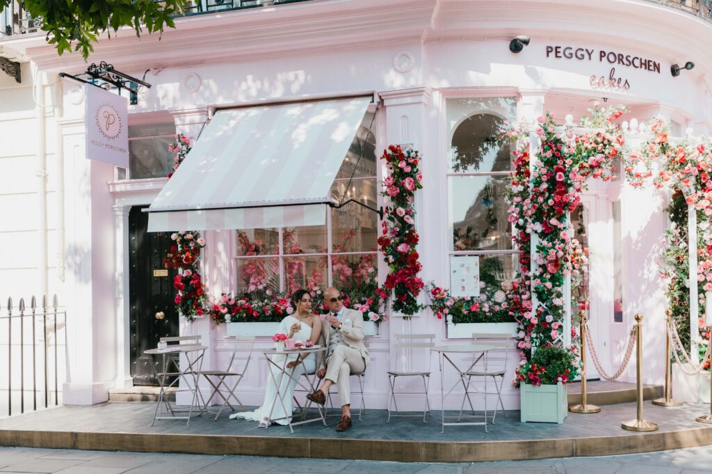 Raissa and Vasileios sit outside Peggy Porschen Café, clinking coffee cups with playful smiles.