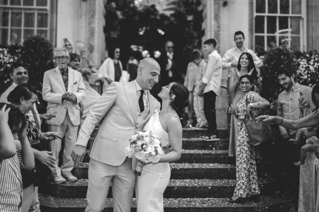 A wide shot of the couple on the steps, with guests cheering and throwing confetti in the background.