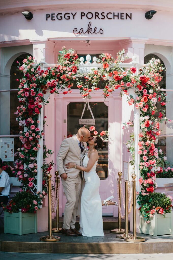 The bride and groom pose in front of Peggy Porschen’s pink storefront, with its signature floral archway adding a romantic backdrop