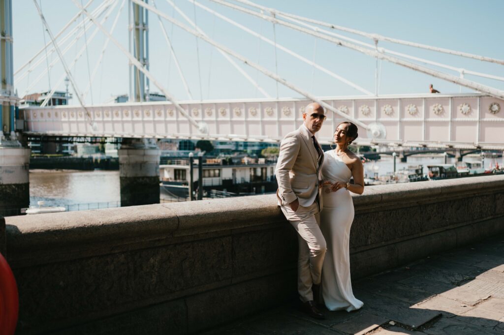 A wide shot of the couple standing on Albert Bridge, with the Thames and greenery visible in the distance.