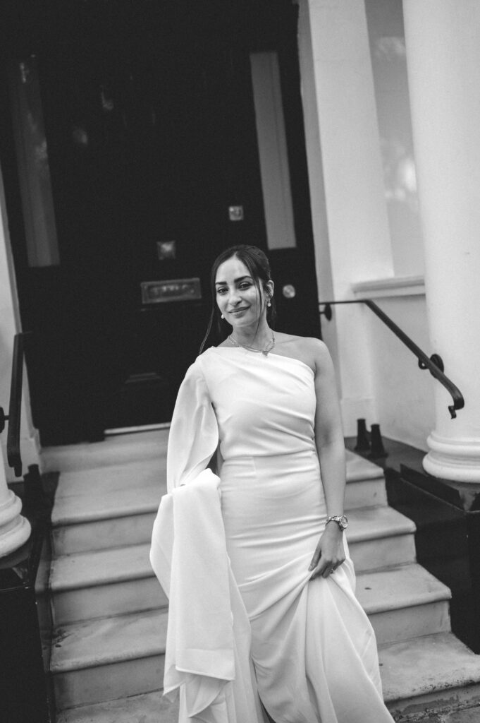 In a timeless black and white frame, Raissa stands elegantly on the steps of a grand townhouse, her minimalist wedding gown flowing beautifully as her serene smile contrasts against the striking black door and classic columns.