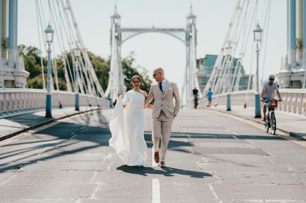 The couple strolls hand-in-hand across Albert Bridge, the iconic white and pastel structure glowing in the summer sunlight.