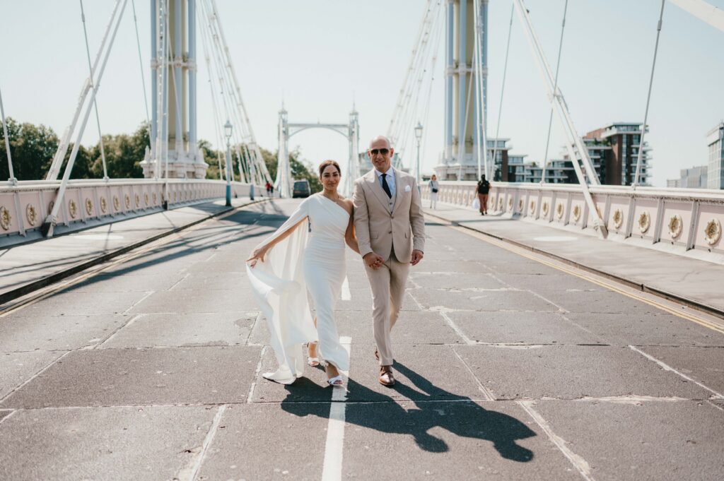 Raissa’s cape sleeve dress flows dramatically as she and Vasileios walk along Albert Bridge, both laughing together.